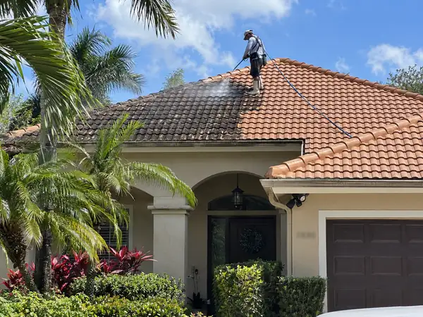 Technician soft-washing a stained terracotta tile roof, with the cleaned half visibly brighter than the dark half